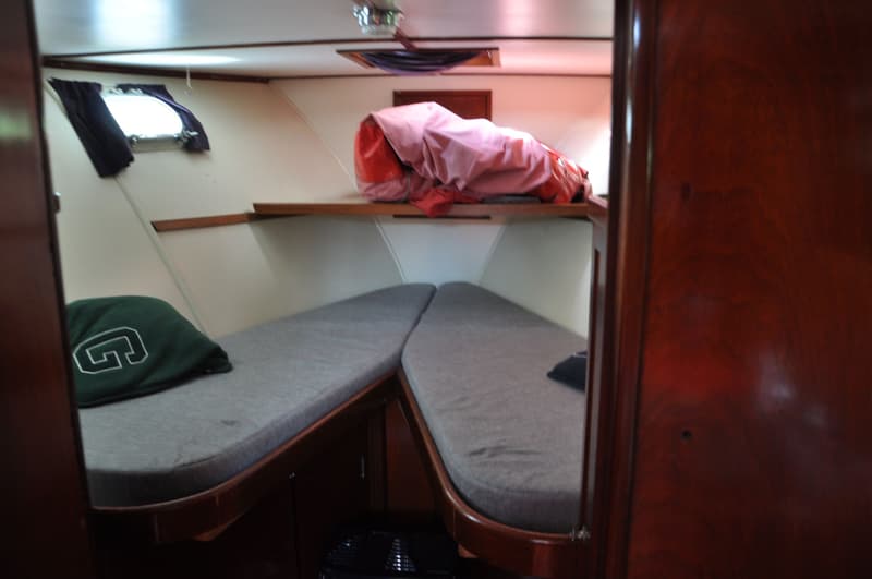 De foto toont een kajuit met twee bedden met grijze matrassen, een raam met gordijnen en een plank met een roze zeil.