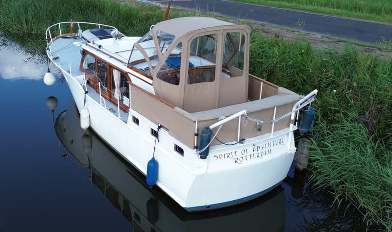 Een witte boot met een beige kap en houten details ligt in het water naast een oever met hoog gras. — Super Van Craft 1170