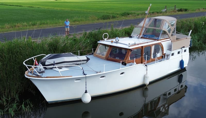 Een witte boot met houten details ligt in het water, met een man op de kant en een groen veld op de achtergrond.