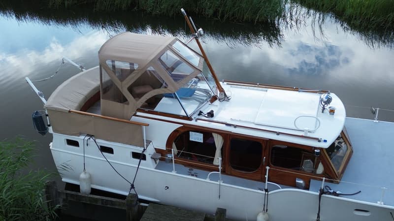 Een witte boot met een bruine kap en houten details ligt aangemeerd aan een steiger, omringd door riet.