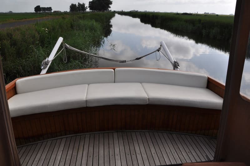 De foto toont het voordek van een boot met witte kussens en een houten vloer, varend op een kanaal omringd door groen.