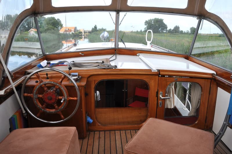 Een stuurhut van een boot met een houten stuurwiel, uitzicht op een waterweg met huizen en groen. — Super Van Craft 1170