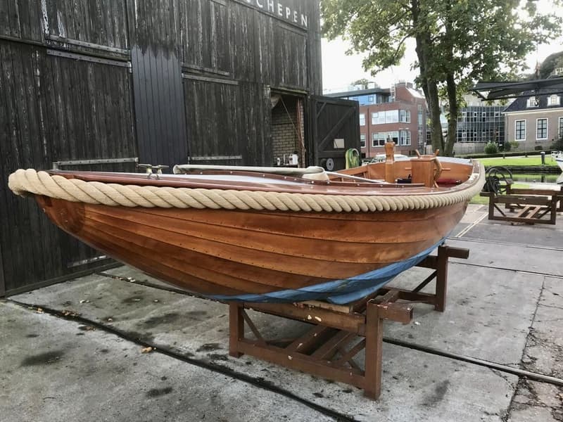 Een houten boot met een touw langs de boeg en een blauwe onderkant, staat op een houten bok voor een donker gebouw.