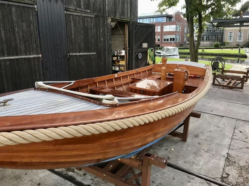 Een houten boot met een touw langs de rand staat op bokken voor een donkerhouten schuur. — De Jong Vlet 6.20