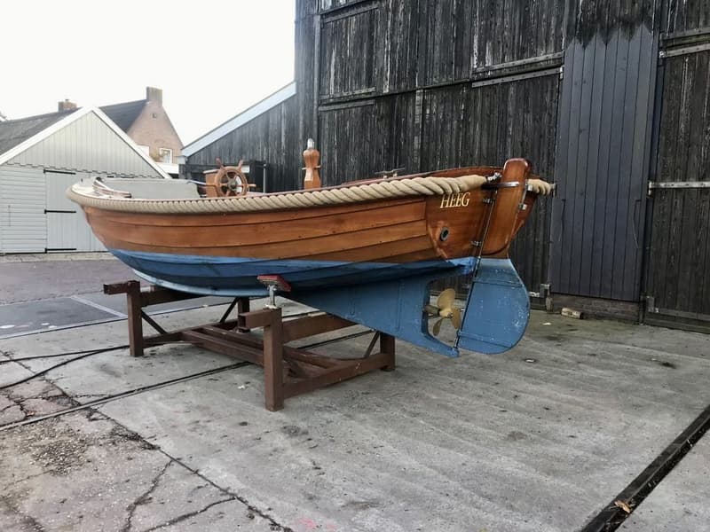 Een houten boot met de naam "HEEG" op de spiegel, staat op een trailer voor een houten schuur. — De Jong Vlet 6.20