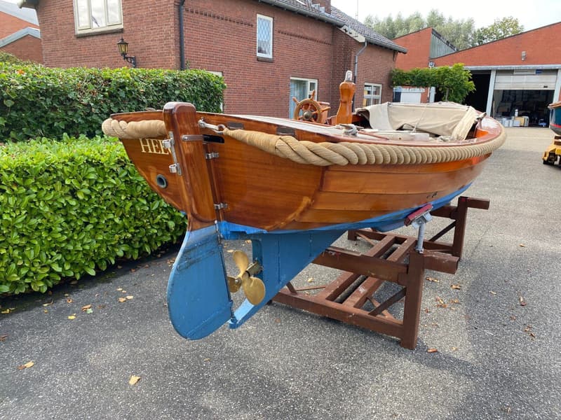 Een houten boot met een blauwe onderkant en een gouden schroef staat op een bruine bok op een geasfalteerde ondergrond.