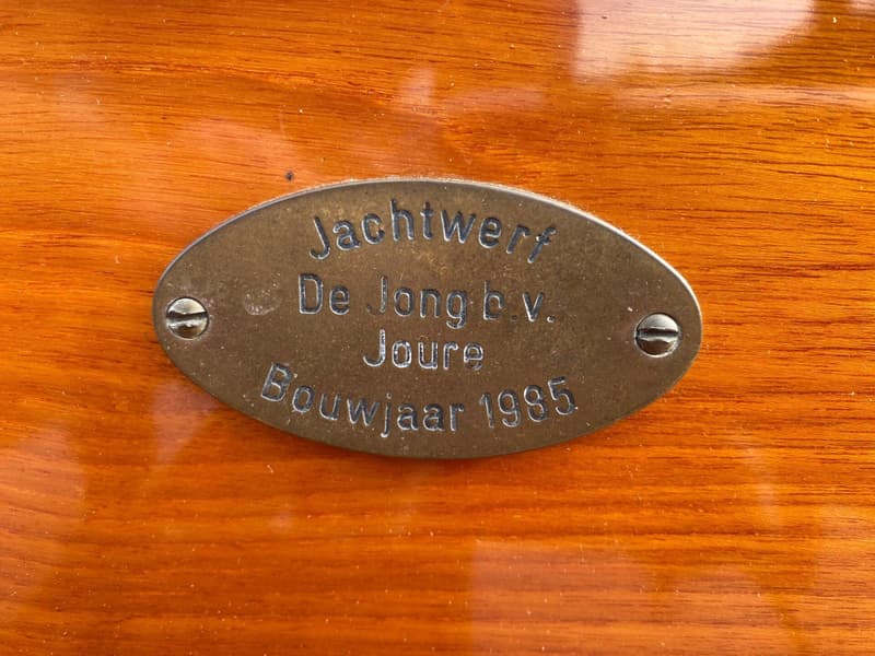 Een ovale, bronzen plaat met de tekst "Jachtwerf De Jong b.v. Joure Bouwjaar 1985" is vastgeschroefd op een houten oppervlak.