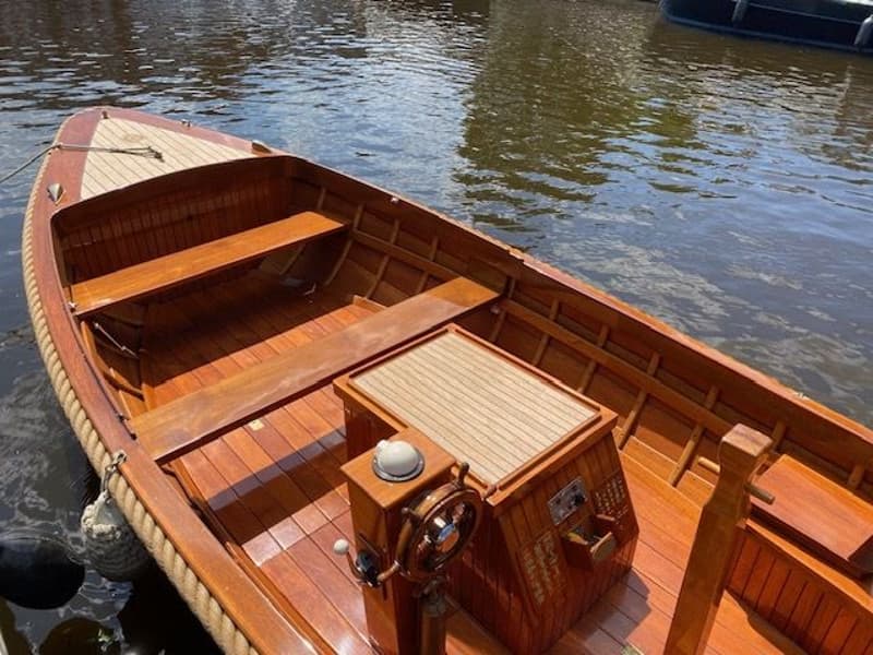 Een houten boot met een stuurwiel, kompas en een touw langs de rand, drijvend in het water. — De Jongvlet 6.20