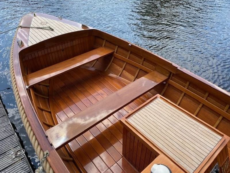 Een houten boot met een touw langs de rand ligt aan een houten steiger, met glanzende houten banken en vloer.