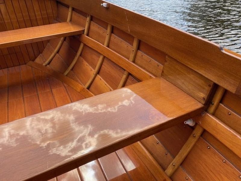 De foto toont het interieur van een houten boot met banken en een glanzende afwerking, met de weerspiegeling van wolken op…