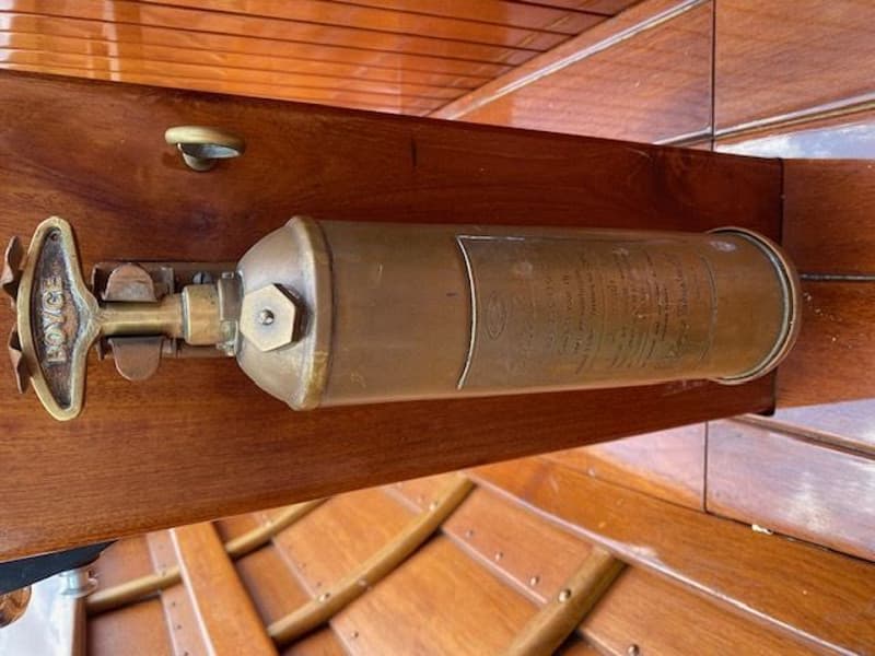 Een koperkleurige brandblusser met het woord "BOYCE" op het handvat, gemonteerd op een houten paneel.