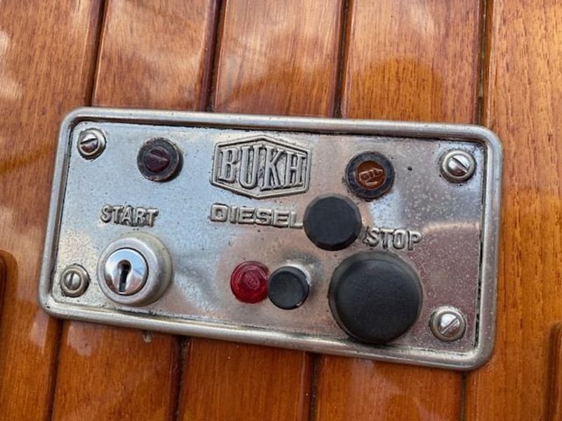 Een zilverkleurig bedieningspaneel met "BUKH DIESEL", "START" en "STOP" knoppen, gemonteerd op een houten oppervlak.