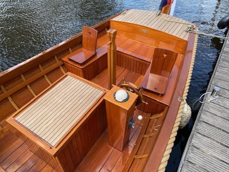 Een houten boot met een stuurwiel, twee stoelen met hartvormige uitsparingen en een tafel, ligt afgemeerd aan een steiger.