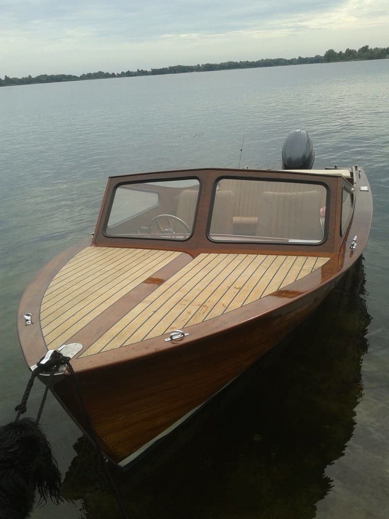 Een houten motorboot met een teakdek en een buitenboordmotor ligt in het water, met een beboste oever op de achtergrond.