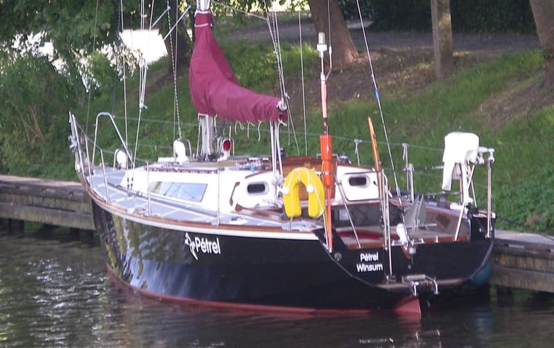 Een zwarte zeilboot met een rode zeilhoes en de naam "Pétrel Winsum" ligt aangemeerd aan een houten steiger.