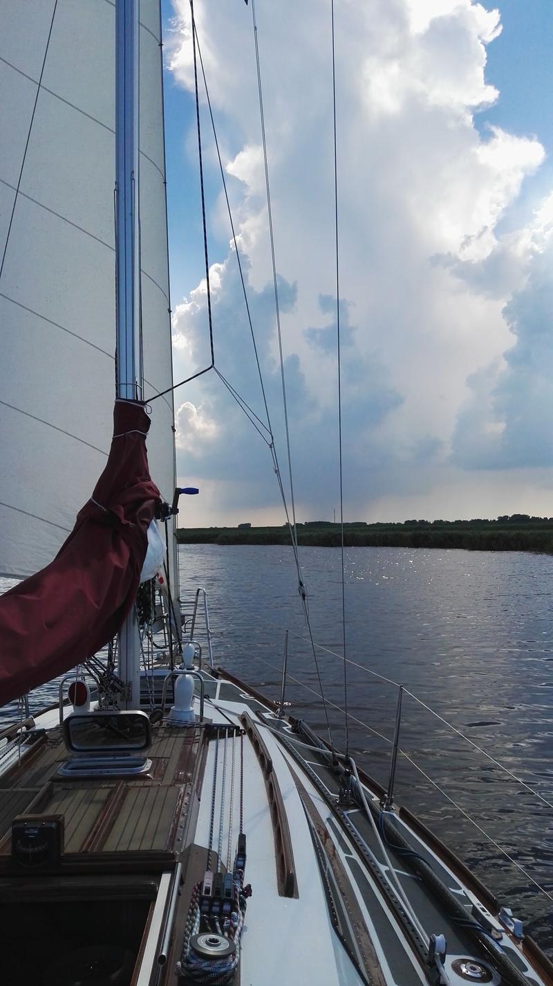Een zeilboot vaart op een waterweg met een bewolkte lucht en een groene oever op de achtergrond. — Waarschip 900 +