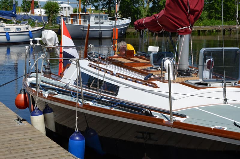 Een witte zeilboot met een rood zeil en een Nederlandse vlag ligt afgemeerd aan een houten steiger. — Waarschip 900 +