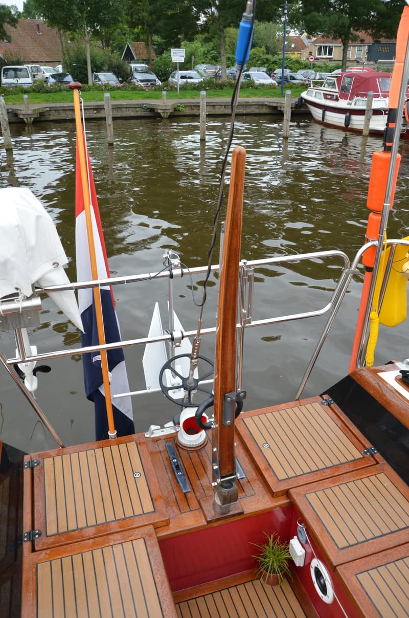 De achterkant van een boot met een Nederlandse vlag, een roer, een kompas en een potplant. — Waarschip 900 +