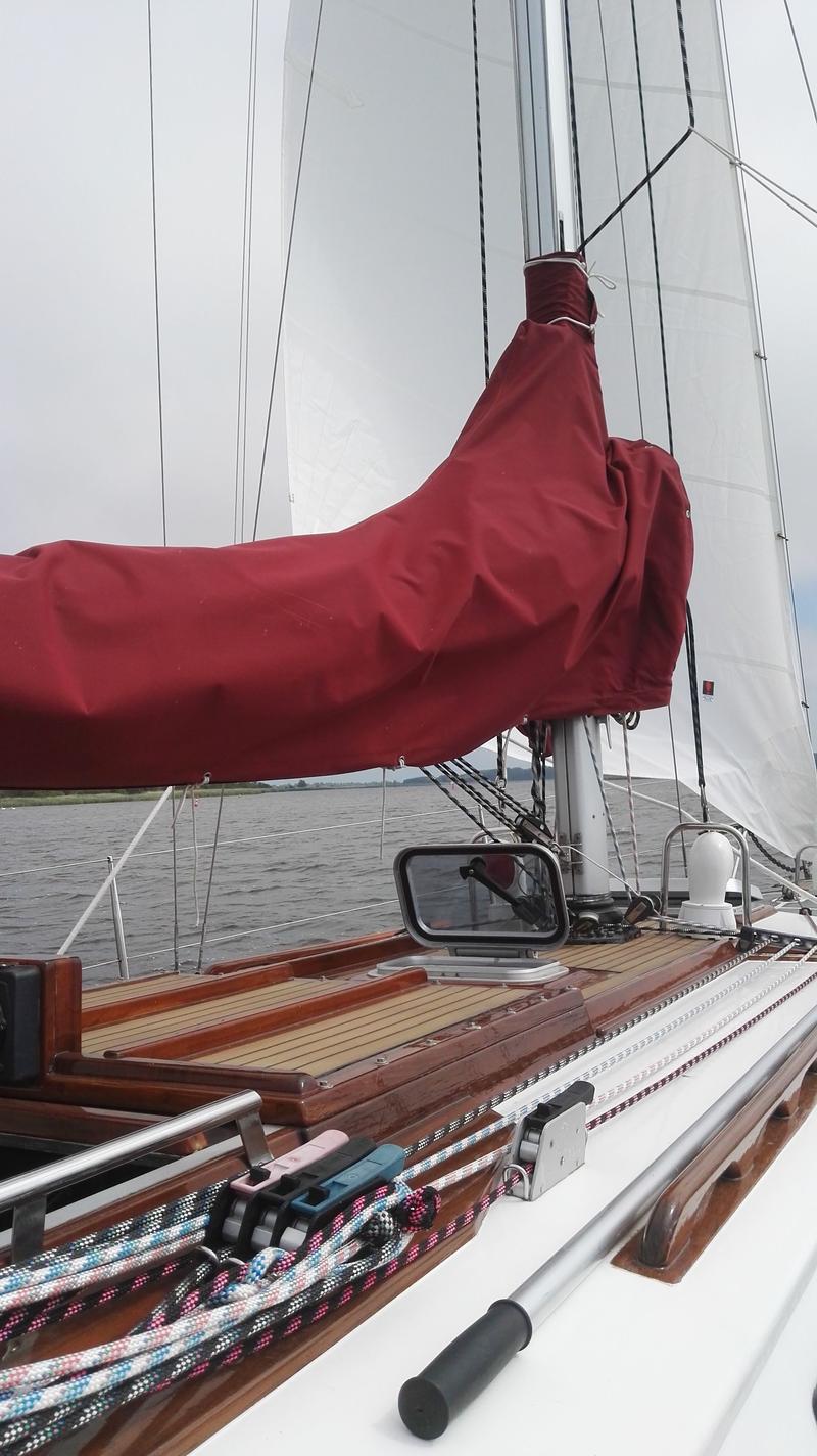 Een zeilboot met een rood zeilhuik, een mast en witte zeilen, varend op water met een landschap op de achtergrond.