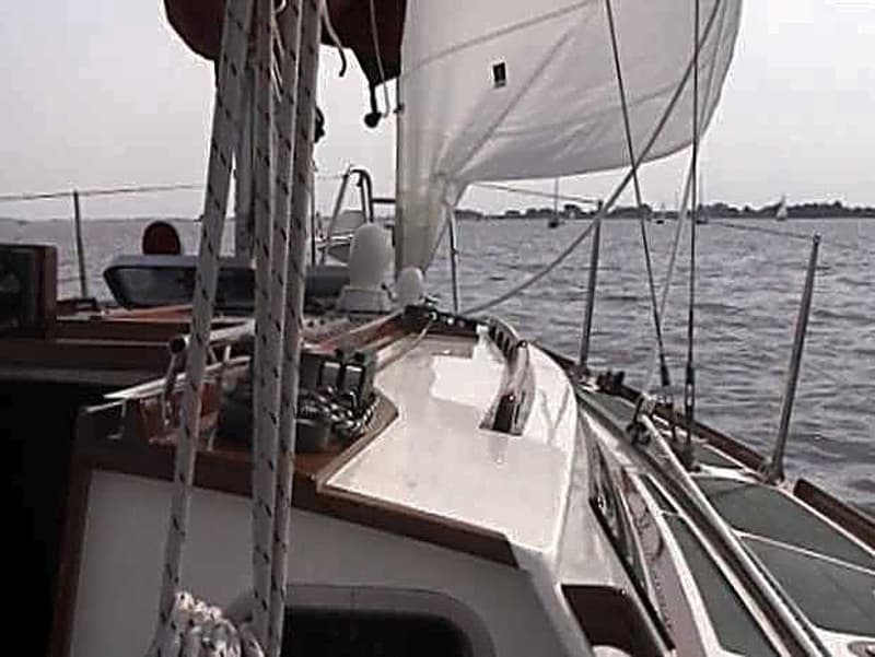 Een zeilboot vaart op water met een wit zeil en touwen zichtbaar, met land en andere boten op de achtergrond.