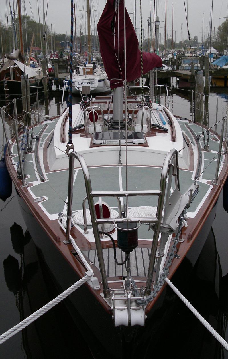 Een zeilboot ligt afgemeerd in een haven, met een donkerrood zeil opgerold aan de mast en een anker aan de boeg.