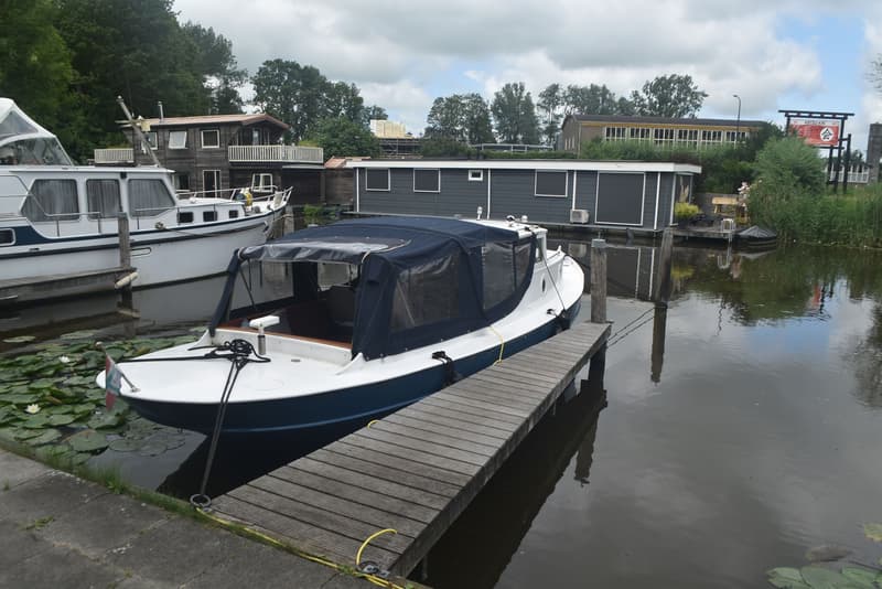 Een witte boot met blauwe romp en donkerblauwe kap ligt afgemeerd aan een houten steiger, omringd door waterlelies.