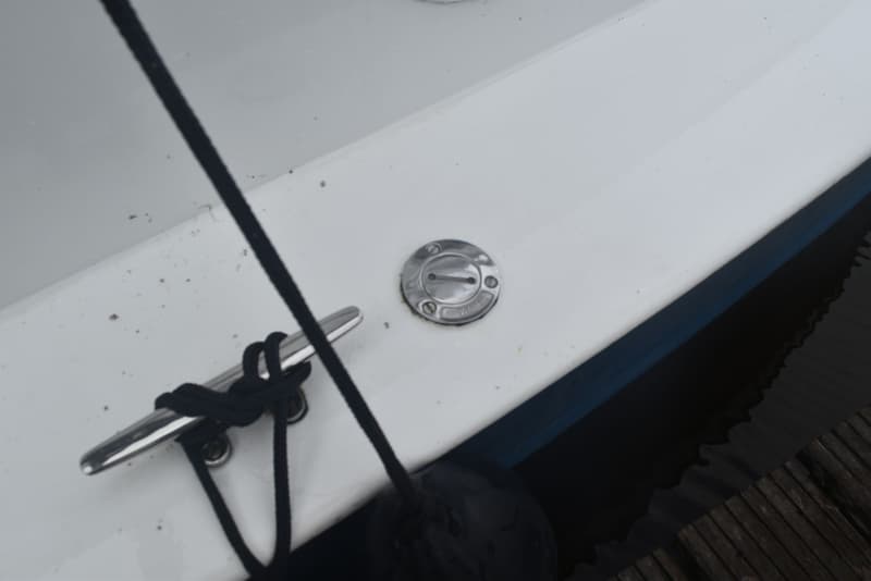 Een witte boot met een zwarte lijn, een zilveren kikker en een zilveren waterdop, naast donker water en een houten steiger.