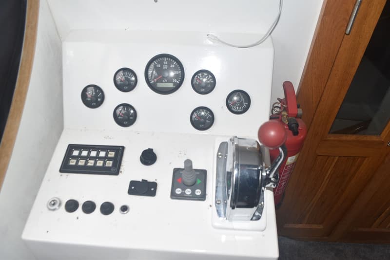 Een wit bedieningspaneel met diverse meters, schakelaars, een joystick en een gashendel, naast een rode brandblusser en een…