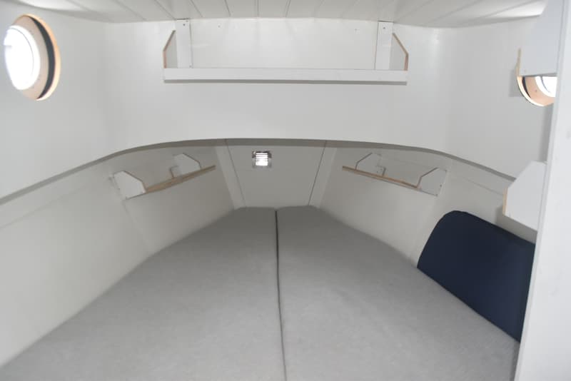 De foto toont een kajuit met twee bedden, twee patrijspoorten, een ventilatierooster en planken aan de zijkanten.