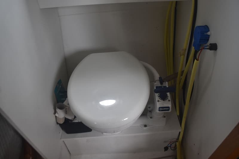 Een wit scheepstoilet met een handpomp en gele slangen is zichtbaar in een kleine, witte ruimte.