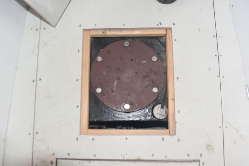 Een ronde, roestbruine metalen plaat met zes zilverkleurige bouten is zichtbaar in een vierkante houten omlijsting.