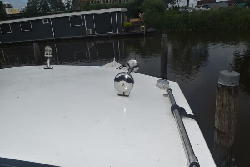 De foto toont het voordek van een boot met een scheepshoorn en een navigatielicht, naast een houten paal in het water.