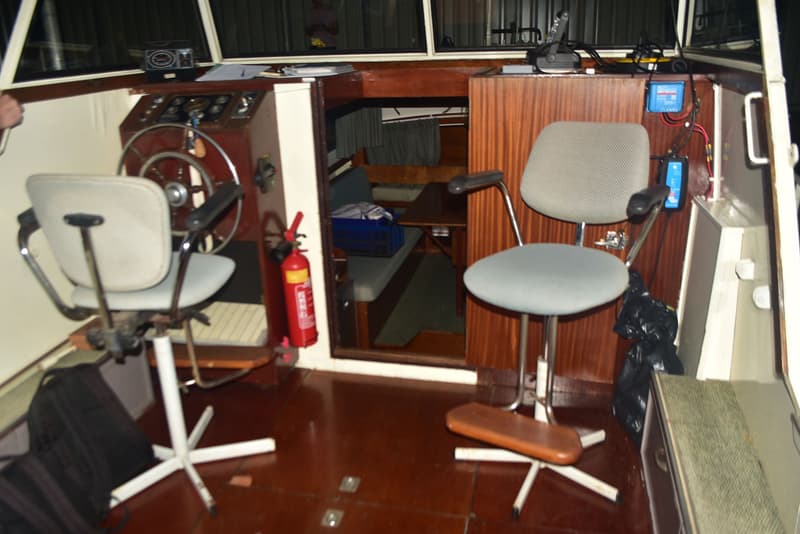 De foto toont het interieur van een boot met een stuurstand, twee stoelen, een brandblusser en een doorgang naar een kajuit.