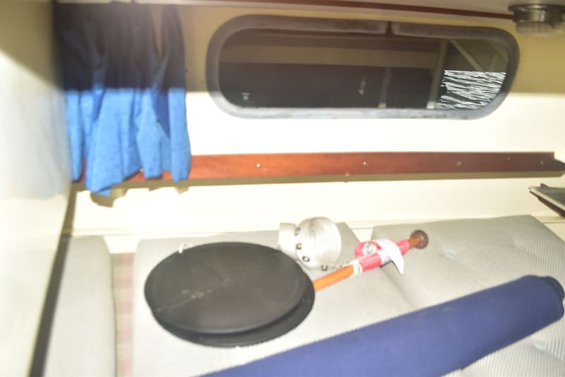 De foto toont een interieur van een boot met een patrijspoort, blauwe gordijnen, een houten plank en diverse voorwerpen op…
