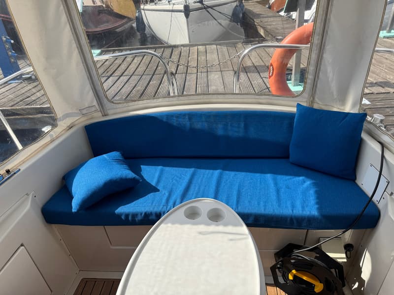 De foto toont een bootinterieur met blauwe kussens en een witte tafel, met uitzicht op een steiger en andere boten.