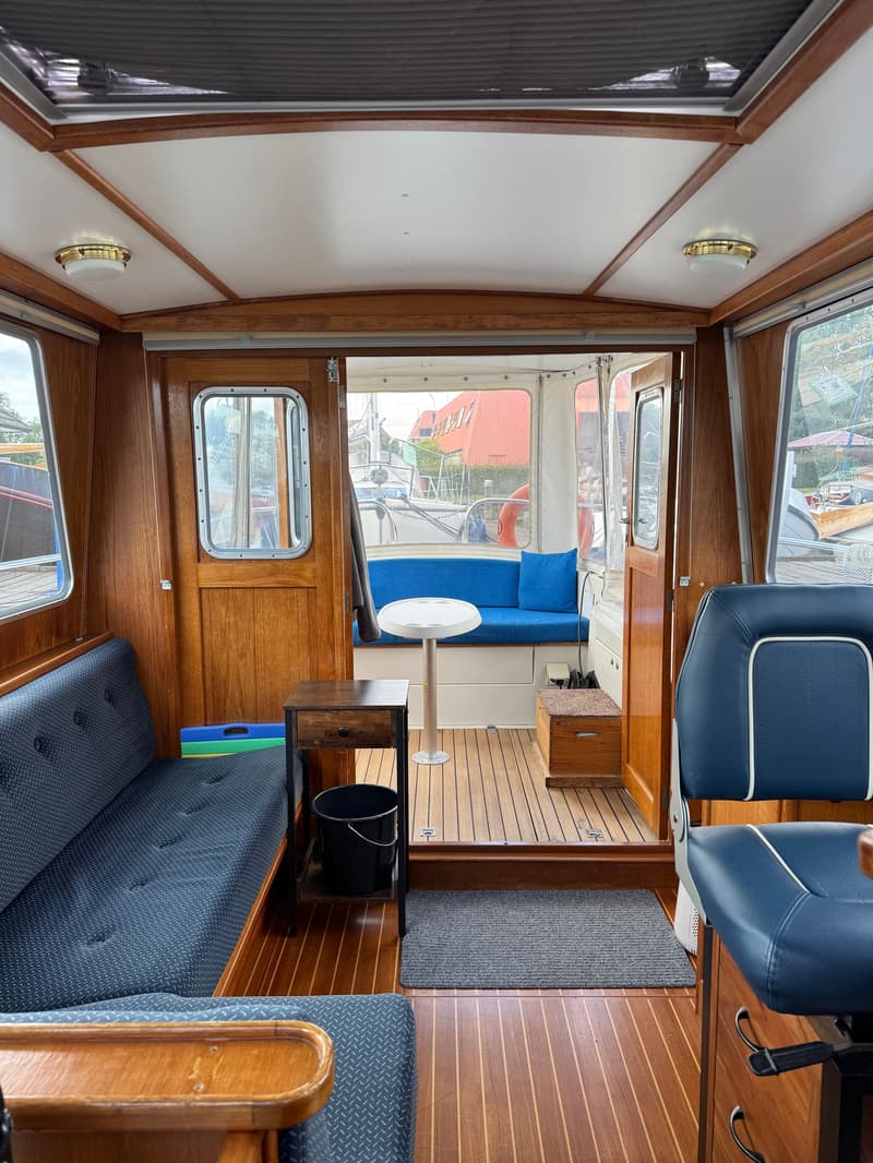 De foto toont het interieur van een boot met houten wanden en vloer, blauwe zitbanken en een stuurstoel.