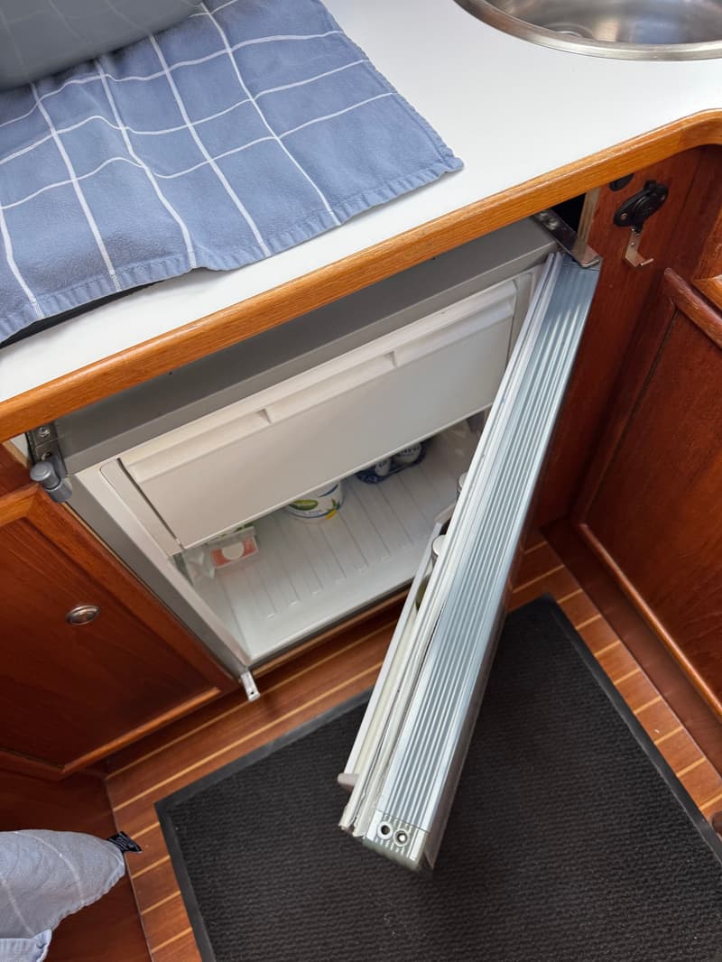 Een open koelkast in een boot met een blauw geruit doek, een gootsteen en een houten interieur. — Drammer 8.20 Classic