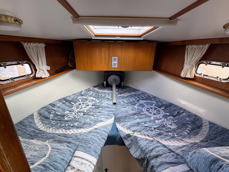 De foto toont de kajuit van een boot met een V-vormig bed, patrijspoorten, een dakluik en houten kasten.
