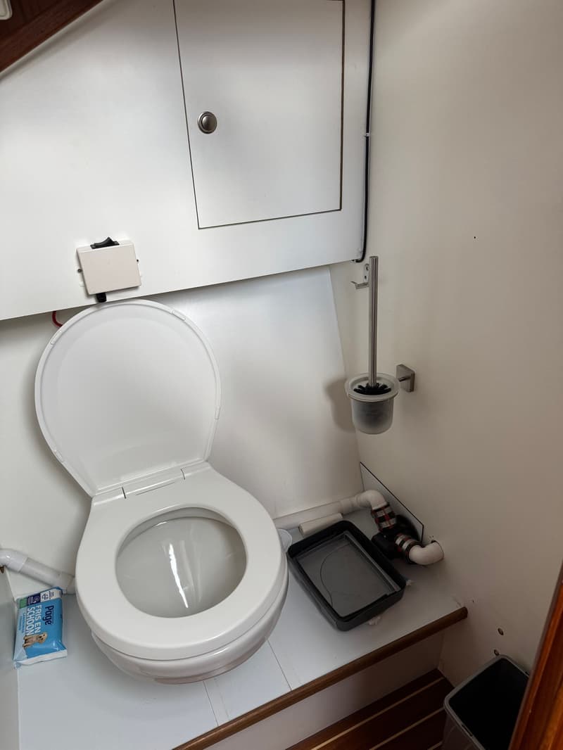 Een wit scheepstoilet met open deksel en zitting, met een toiletborstel en een pakje vochtige doekjes ernaast.