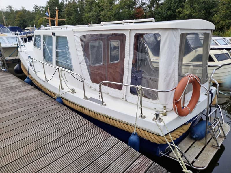 Een witte boot met een blauwe romp ligt afgemeerd aan een houten steiger, met een reddingsboei aan de achterkant.