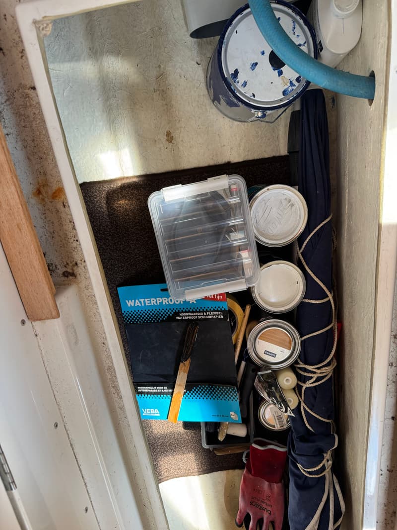 Een opslagruimte met verfblikken, schuurpapier, een opgerolde paraplu, een touw en een werkhandschoen.