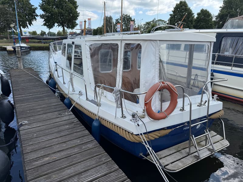 Een witte boot met blauwe romp en een bruine stootrand ligt afgemeerd aan een houten steiger. — Drammer 8.20 Classic