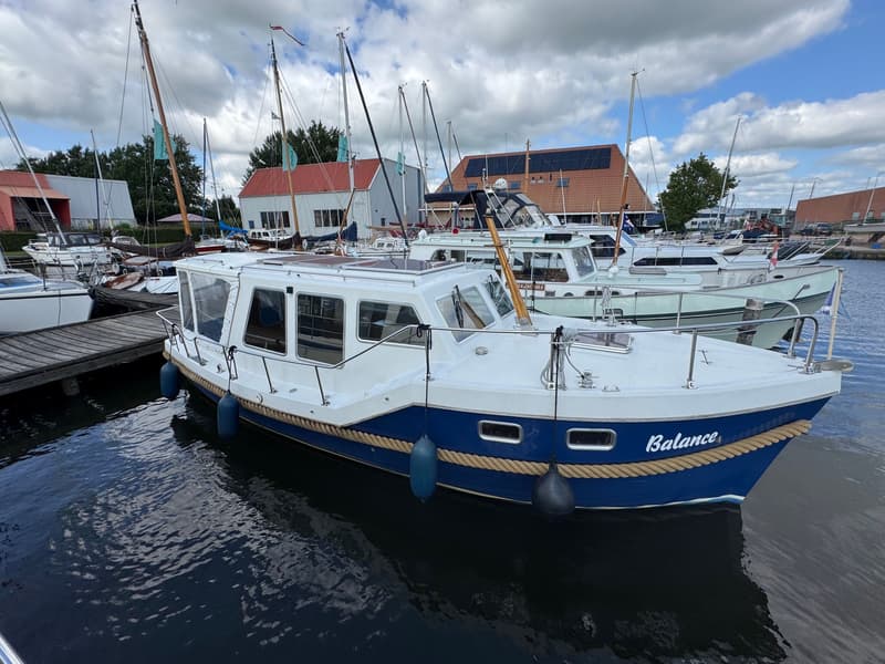 Een witte en blauwe motorboot met de naam "Balance" ligt aangemeerd in een haven, omringd door andere boten en gebouwen.