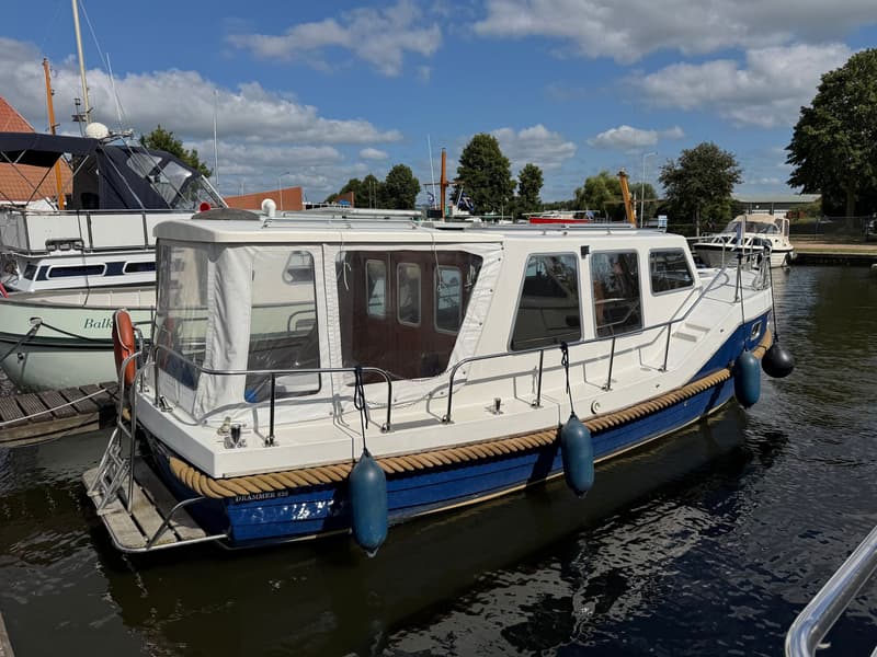 Een witte boot met blauwe romp en een bruine stootrand ligt in het water, met de naam "DRAMMER 820" op de achtersteven.