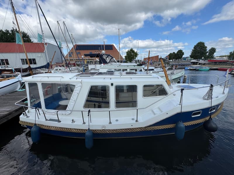 Een witte boot met een blauwe romp ligt afgemeerd in een haven, met andere boten en gebouwen op de achtergrond.