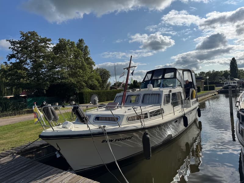 Een witte motorboot met de naam "BON VOYAGE" ligt afgemeerd aan een houten steiger onder een blauwe lucht met wolken.