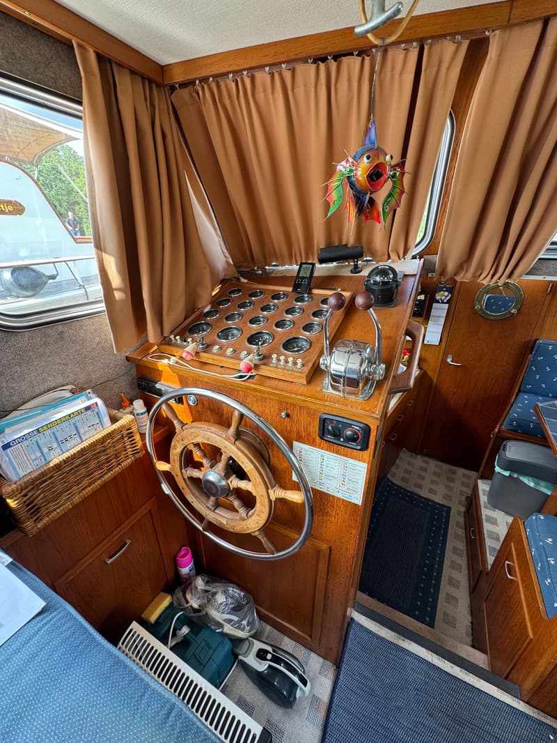 De foto toont het interieur van een boot met een houten stuurwiel, een dashboard vol meters en bedieningshendels.