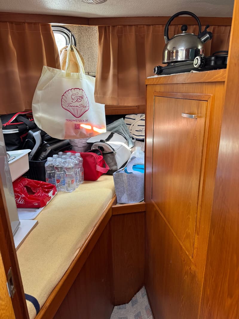De foto toont een interieur met houten wanden, een raam met bruine gordijnen, een waterkoker op een kookplaat en diverse…