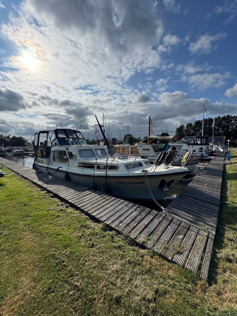 Een witte motorboot met de naam "BON VOYAGE" ligt aangemeerd aan een houten steiger, met gras ernaast. — Neptunus 107 TWIN