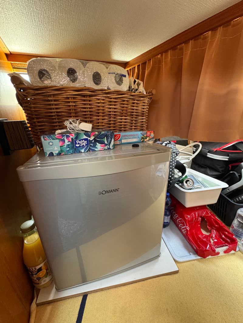Een zilverkleurige Bomann koelkast staat op een houten plank, met een fles sap ernaast en een rieten mand erboven.